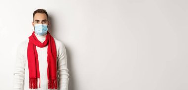 Covid-19, tecrit ve karantina konsepti. Beyaz arka planda duran, kışın giydiği süveter ve sağlık maskeli genç adam..