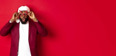 Komik siyah adam yeni yılı kutluyor, parti gözlüğü takıyor ve Noel Baba şapkası takıyor, kırmızı arka planda eğleniyor..