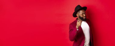 Şüpheli siyah adam kamerayı işaret ediyor ve gözlerini kısıyor, seni suçluyor, uyarı veriyor, kırmızı arka planda duruyor..