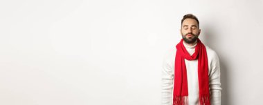 Kapalı gözleri ve büzüşmüş dudakları olan, ökseotunun altında Noel öpücüğü bekleyen, beyaz arka planda duran, kışın atkı ve süveteri olan yakışıklı adam..