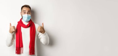 Covid-19, tecrit ve karantina konsepti. Yüz maskesi takmış süveter ve kırmızı eşarplı, beyaz arka planda baş parmağını gösteren bir adam..