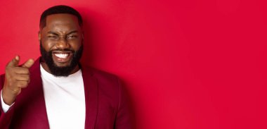 Sakallı neşeli siyah adam, parti kıyafetleri giyiyor, kamerayı işaret ediyor ve göz kırpıyor, yılbaşı ve yılbaşı ile kutluyor, kırmızı arka planda duruyor..