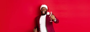 Noel, parti ve tatil konsepti. Yakışıklı siyah adam Noel Baba şapkalı, şampanya kaldıran, gülümseyen, kadeh kaldıran, yeni yılı kutlayan, kırmızı arka planlı.