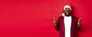 Komik siyah adam yeni yılı kutluyor, parti gözlüğü ve Noel Baba şapkası takıyor, başparmağını kaldırıyor, onaylıyor ve kırmızı arka planda duruyor..