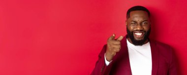 Sakallı neşeli siyah adam, parti kıyafetleri giyiyor, kamerayı işaret ediyor ve göz kırpıyor, yılbaşı ve yılbaşı ile kutluyor, kırmızı arka planda duruyor..