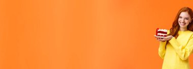 Sevimli kızıl saçlı kadın elinde lezzetli ev yapımı pastalar tutuyor ve gülümsüyor, ısırıyor, ısırıyor, arkadaşlarına davranıyor, doğum gününü kutluyor gibi sırıtıyor ya da lezzetli tatlı tatlılar yiyor gibi, turuncu arka plan.