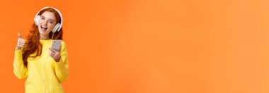 Çekici kızıl saçlı kız yeni bir Noel hediyesi aldı. Havalı kulaklıklar, kulaklıkla müzik dinledi ve başparmağını kaldırdı, akıllı telefon tuttu, müzikten hoşlandı, şarkı söyledi ve gülümsedi, turuncu arka plan..