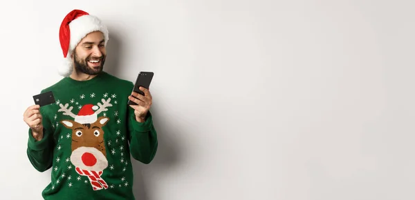 オンラインショッピングやクリスマスのコンセプト 幸せな髭を生やした男がインターネットでクレジットカードと電話で購入し サンタの帽子とセーターを着て 白い背景 — ストック写真