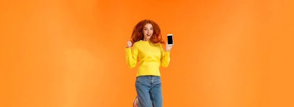Advertising, technology and holidays concept. Vertical full-length shot cheerful gorgeous ginger girl, redhead woman jumping over orange background with smartphone, show thumb-up.