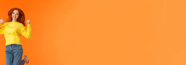 Shopping, fashion and women concept. Vertical full-length positive and cute redhead young modern hipster girl in denim jeans, winter yellow sweater jumping and showing thumbs-up in approval, smiling.