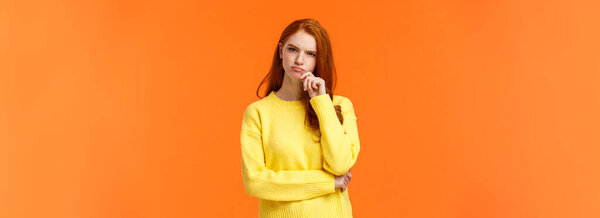 Skeptical and unsure redhead focused, serious-looking female taking hard decision, making choice gift make winter holidays, looking shopping aisle, touch lip consider, thinking, orange background.