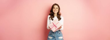 Celebration and holidays. Happy beautiful girl holding gift, wrapped present and smiling, receive surprise, standing against pink background.