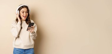 Smiling happy woman listening music in headphones on smartphone, laughing excited, standing over beige background.