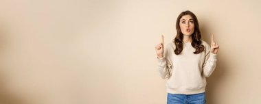 Amazed girl checking out banner announcement, reading promo text upwards, pointing fingers up, standing over beige background.