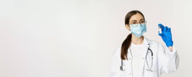 Genç kadın portresi, tıbbi maske ve üniformalı doktor, aşıyı gösteriyor, covid-19 aşı kampanyası, beyaz arka planda duruyor..