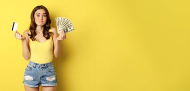 Young pretty woman in summer outfit holding credit card and money, cash in hands, standing against yellow background.