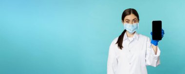 Young woman doctor in medical face mask and hospital uniform, showing mobile phone app, screen interface, online clinic concept, standing over torquoise background.