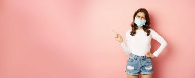 Confused, indecisive young woman in medical face mask, pointing finger left and looking complicated, making decision, standing over pink background.