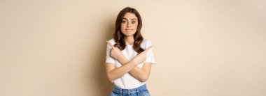 Beautiful girl choosing between two options, pointing sideways and looking clueless, standing over beige background.