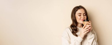 Woman smiling and smelling delicious cup of coffee in takeaway cup, standing over beige background.