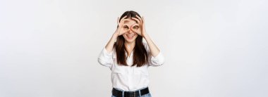 Funny girl looking through hand binoculars, zero gesture, standing over white background and smiling, seeing smth interesting far away, white background.