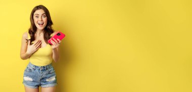 Surprised smiling woman holding mobile phone, reacting amazed and impressed at camera, seeing something awesome, standing against yellow background.