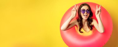 Portrait of happy stylish girl in sunglasses, inside beach swimming ring, showing peace sign, laughing and smiling, standing over yellow background.