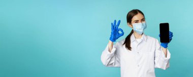 Portrait of doctor in medical face mask and gloves, showing mobile phone app, smartphone screen and okay sign, recommending online checkup website, standing over torquoise background.