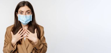 Workaplce ve pandemik konsept. Şok olmuş tıbbi maskeli iş kadını, soluk soluğa, kameraya korkmuş ve endişeli bakıyor, beyaz arka planda duruyor..