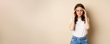 Health and women concept. Portrait of girl feeling sick, touching head temples, feeling headache, migraine, standing over beige background.