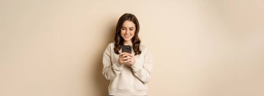 People and cellular technology. Beautiful stylish woman using mobile phone, smartphone app, smiling and looking at screen, standing over beige background.