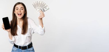 Enthusiastic young woman winning money, showing smartphone app interface and cash, microcredit, prize concept, standing over white background.