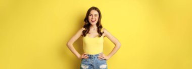 Stylish young brunette woman showing her white healthy teeth, smiling and showing tongue, winking enthusiastic, standing over yellow background.