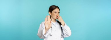 Woman doctor dislike, shut nose from bad smell, reek, showing rejection, stop gesture, standing against torquoise background.