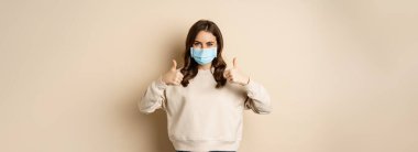 Covid-19, salgın ve karantina konsepti. Genç kadın Coronavirus omicron salgını sırasında tıbbi yüz maskesi takıyor, baş parmaklarını gösteriyor, bej arka planda duruyor..