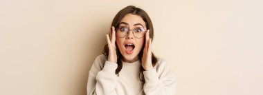 Close up portrait of cute woman in glasses looking impressed, reacting amazed at smth she sees, standing over beige background.