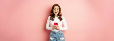 Online shopping and cellphone concept. Excited attractive girl holding mobile phone and smiling, standing over pink background.