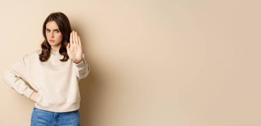 Stop. Serious and confident woman showing extended arm palm, prohibit, forbid smth, blocking something, standing over beige background.