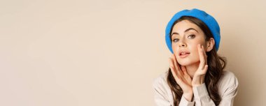 Close up portrait of young beatufiul woman looking in mirror and fixing her makeup, touching soft clear skin, standing over beige background.