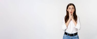 Surprised brunette woman looking amazed, seeing smth awesome, standing in blouse and jeans over white background.