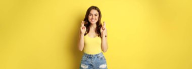 Hopeful young beautiful woman praying, believe, hoping to receive smth, cross fingers for good luck, anticipating, standing over yellow background.