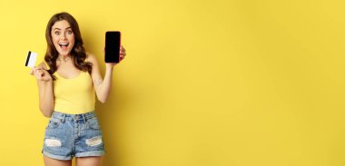 Enthusiastic modern woman showing smartphone screen, holding credit card and smiling, recommending mobile app, standing over yellow background.