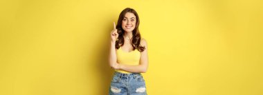 Creative young woman has suggestion, raising finger, eureka sign, has an idea, standing in summer outfit over yellow background. Copy space