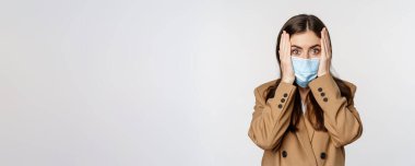 Covid-19 and people concept. Concerned, troubled office lady in medical face mask, holding hands on head and staring shocked at camera, white background.