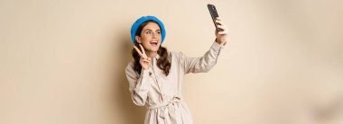Beautiful modern woman posing for selfie, holding smartphone, taking picture photo on mobile phone, standing happy against beige background.
