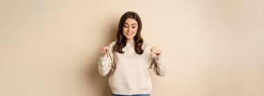 Excited adult woman pointing fingers down and smiling, looking at banner below, showing advertisement, found smth, standing over beige background.