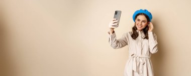 Beautiful girl taking selfie on smartphone in coat and cute hat, smiling for photo and looking happy, standing over beige background.