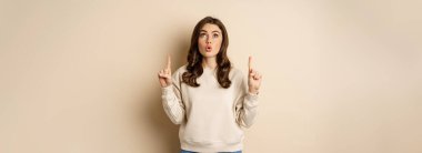 Amazed girl checking out banner announcement, reading promo text upwards, pointing fingers up, standing over beige background.