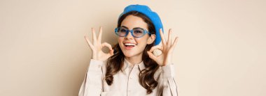 Image of happy smiling woman in stylish sunglasses, showing okay signs in approval, recommending eyewear store, beige background.