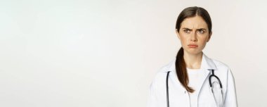 Angry and upset young woman doctor, female healthcare worker sulking, frowning disappointed, dislike smth, standing over white background.